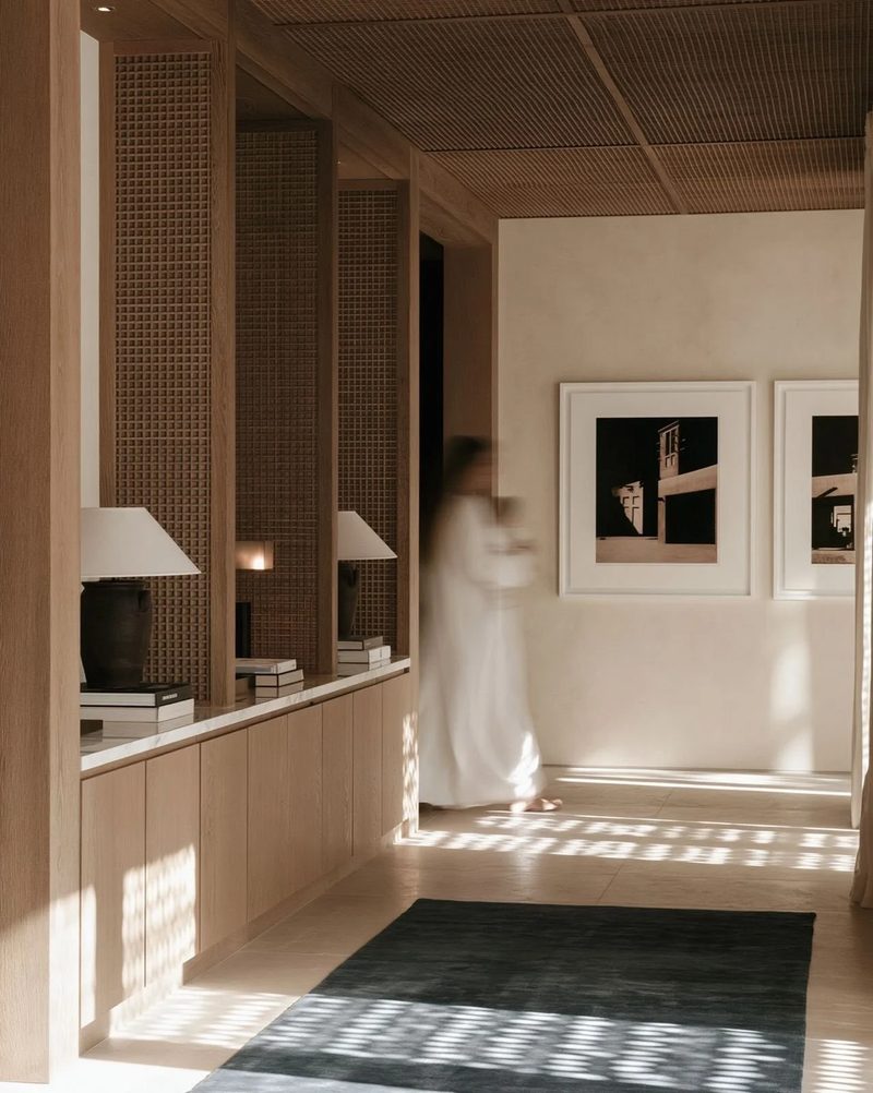 M19 · Árdh, concept image — a calm interior of timber screens and framed photographs, a dark rug catching patterned sunlight, a figure in white by the wall