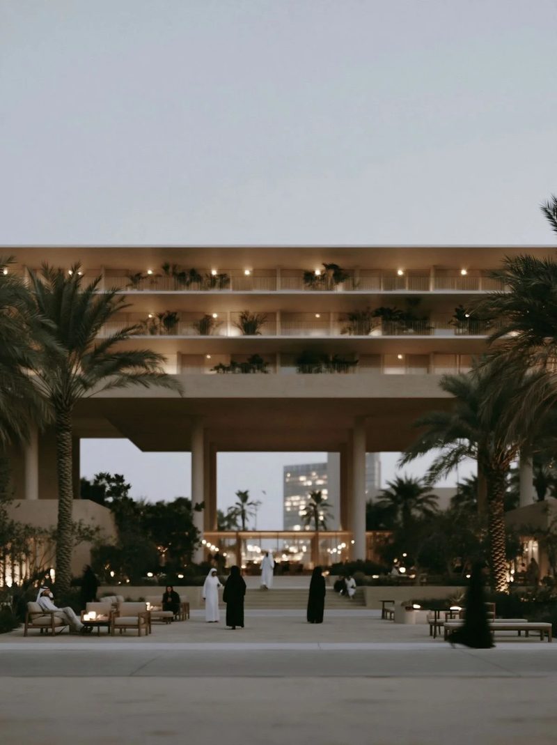 M4 · Árdh, concept image — a deep shaded colonnade under terraced floors at dusk, people sitting among palms